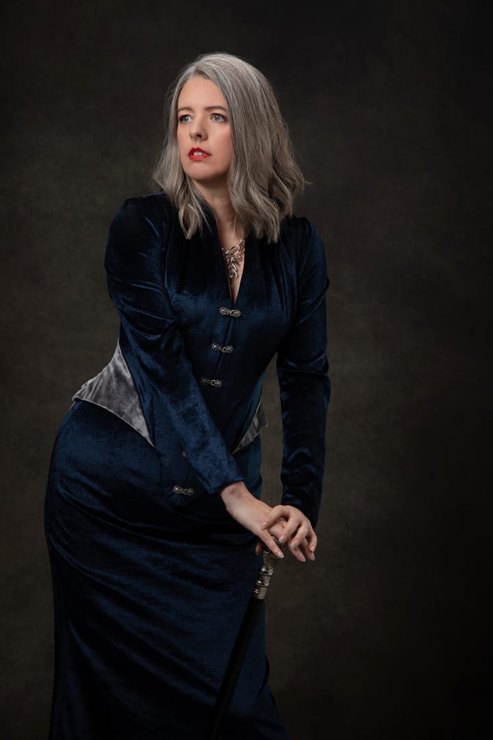 Author Emma Newman wearing a silver and blue velvet outfit and posing with a cane