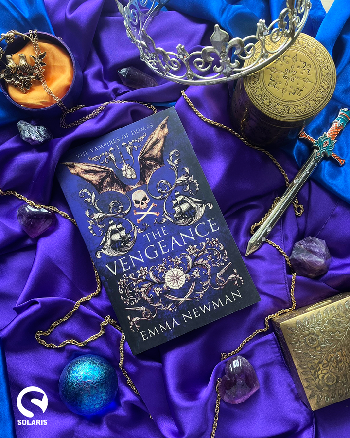 A copy of the novel The Vengeance by Emma Newman, resting on blue silk and surrounded by treasure